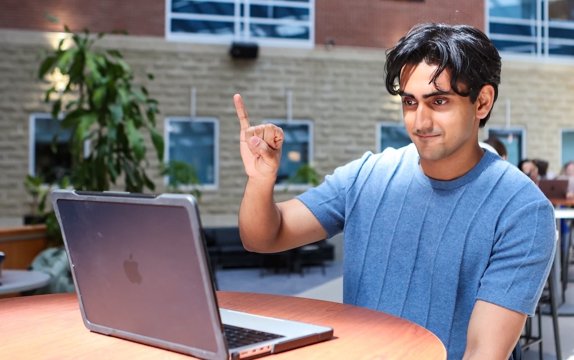 A person in a blue sweater sits a table in an atrium using handshapes to sign in front of a gray laptop