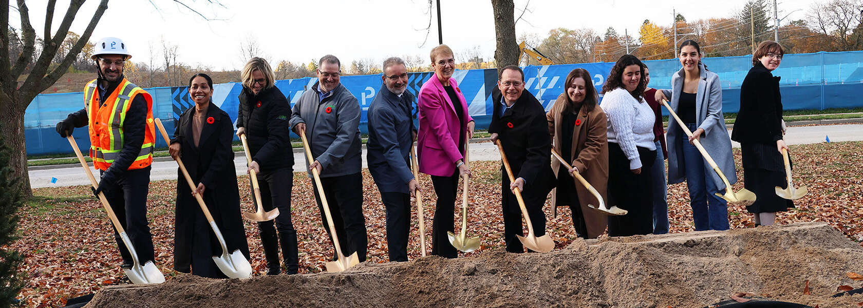 A dozen people use ceremonial shovels in front of a long pile of dirt