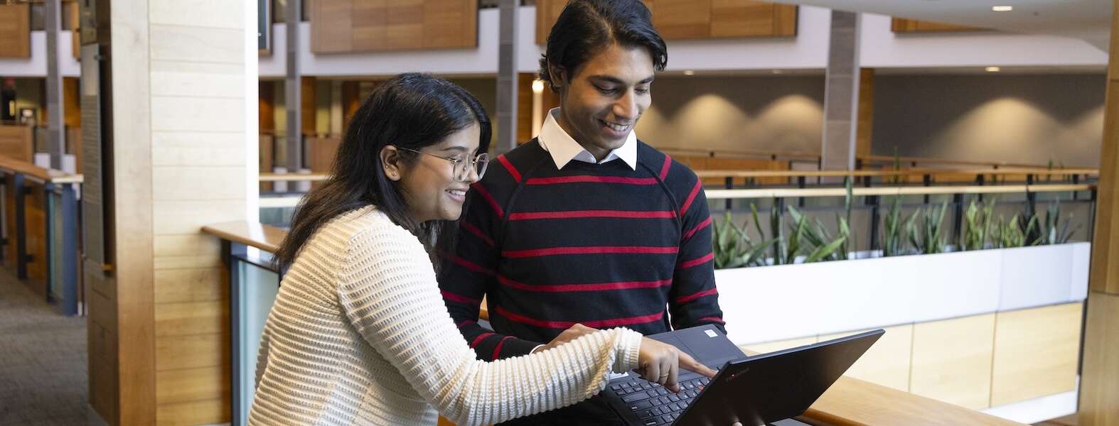 Two people stand together looking at the laptop one is holding in a learning space with plants and wooden walls.