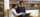Two people stand together looking at the laptop one is holding in a learning space with plants and wooden walls.
