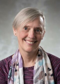 A person with short gray hair wearing a purple, teal, brown and white scarf over a purple shirt smiles into the camera in front of a gray background.