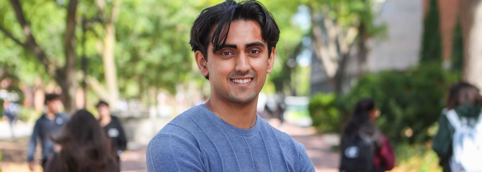 A person in a blue sweater stands smiling into the camera on a walkway surrounded by students and trees on U of G's campus.