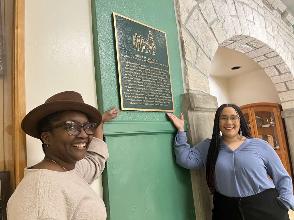Two Black women stand pointing to a green and golden plaque depicting text hung on a green wall.