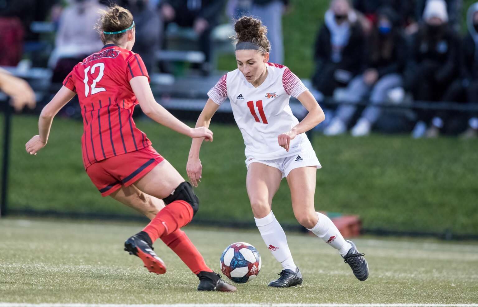 Two soccer players, one in a red uniform and one in a white uniform, run toward each other on a green turf field heading for the ball as spectators watch in the background.
