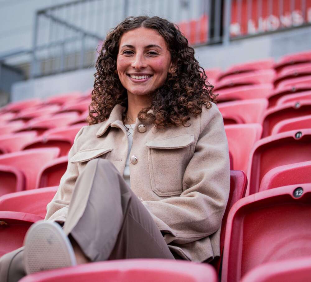 A woman with curly brown hair wearing a brown and beige jacket and brown pants sits in a stadium of red seats smiling into the camera with one leg up.