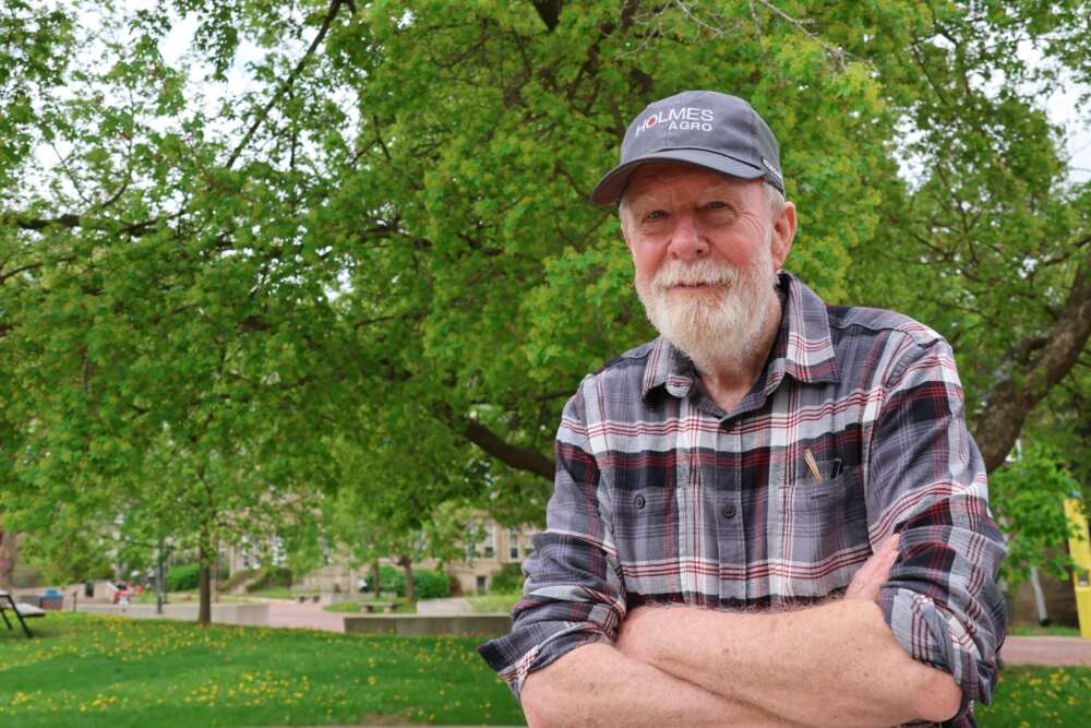 A man with a gray beard in a plaid shirt and hat stands with arms crossed in front of a concrete bench and trees in Branion Plaza.