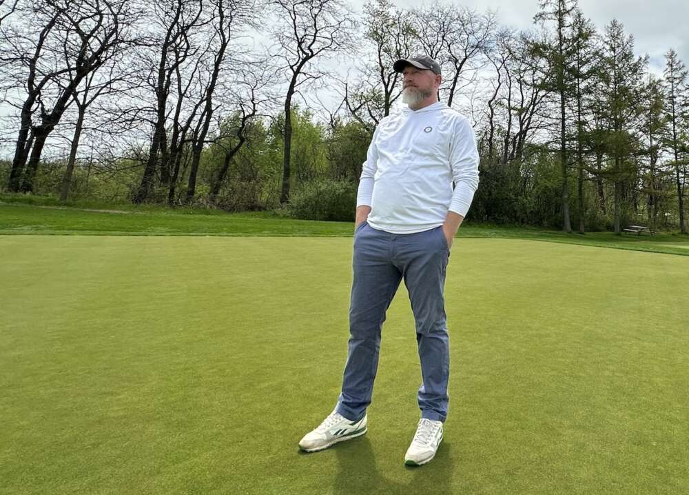A person with a beard wearing white shoes, blue pants, a white hoodie and a blue hat stands in the centre of a field of turfgrass with trees behind them.