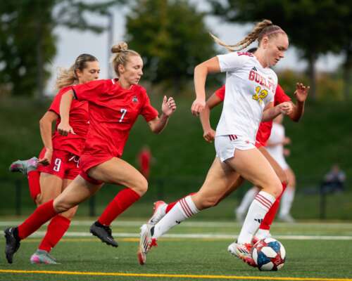 Three soccer players in red uniforms chase one player in a white uniform who has the ball on a green soccer field.