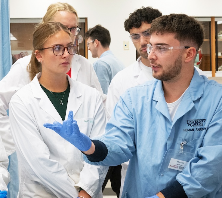 A group of students in white lab coats, goggles and blue latex gloves stand listening to an instructor in a blue lab coat and goggles. 