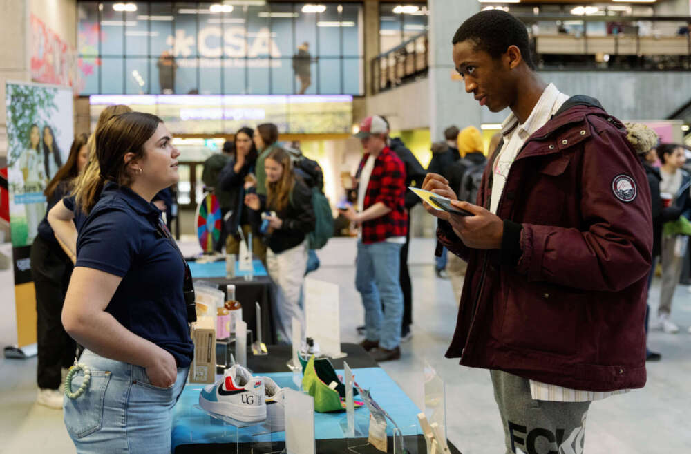 A person wearing a navy blue golf shirt and jeans stands in front of a table of products in the UC speaking to another person in a maroon winter coat holding a cell phone.