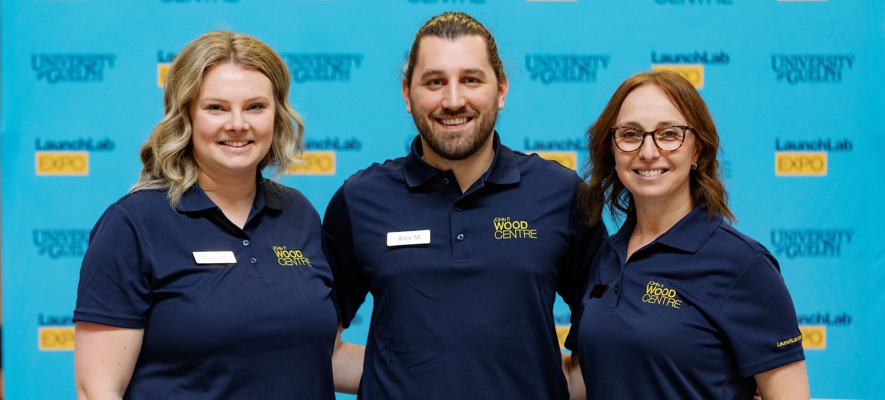 Three people dressed in navy blue Wood Centre golf shirts stand together smiling into the camera with a blue, yellow and black background of text reading University of Guelph and LaunchLab Expo.