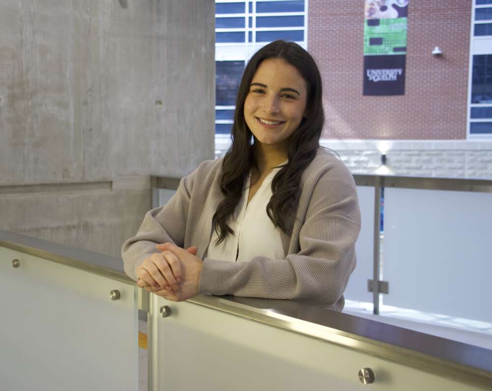 A person with long brown hair wearing a white blouse and gray cardigan clasps her hands while standing over the rail of a staircase in the Waasamowin atrium with red brick and a U of G banner in the background.