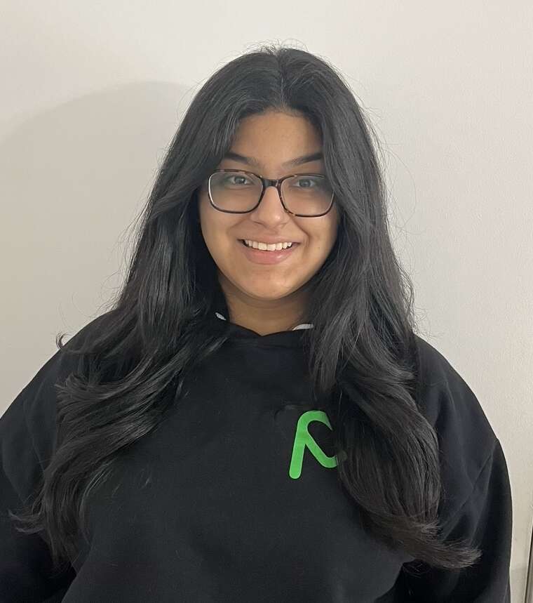 A person with long, brown hair and glasses stands in a black sweatshirt with a green MealCare logo smiling.