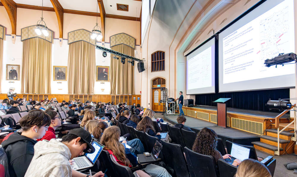 students sit in theatre style seats while a person stands on a platform and addresses them