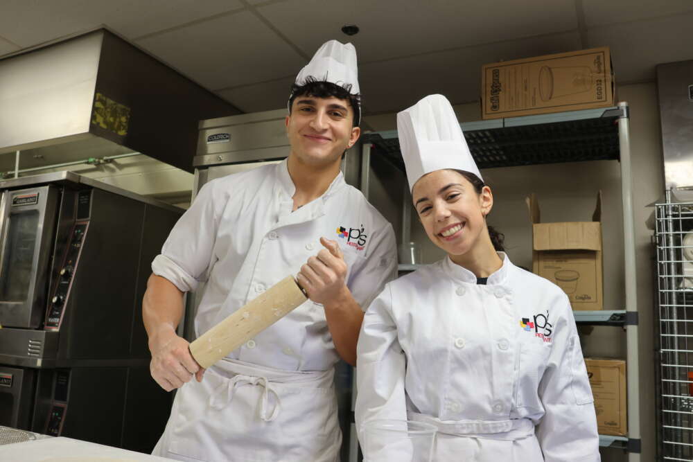 Two students in chef's kitchen whites stand together smiling, one holding a rolling pin.