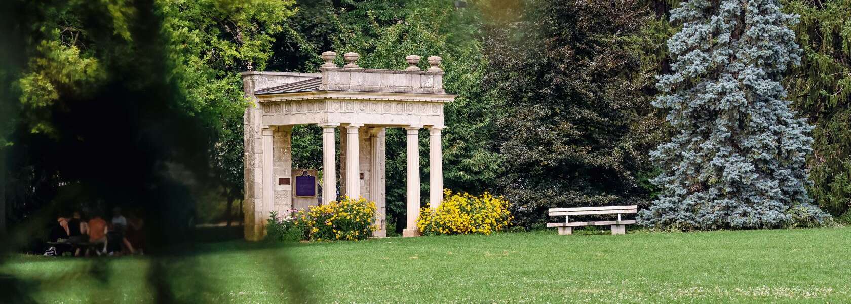 a cream coloured pillared structure on green grass in front of trees