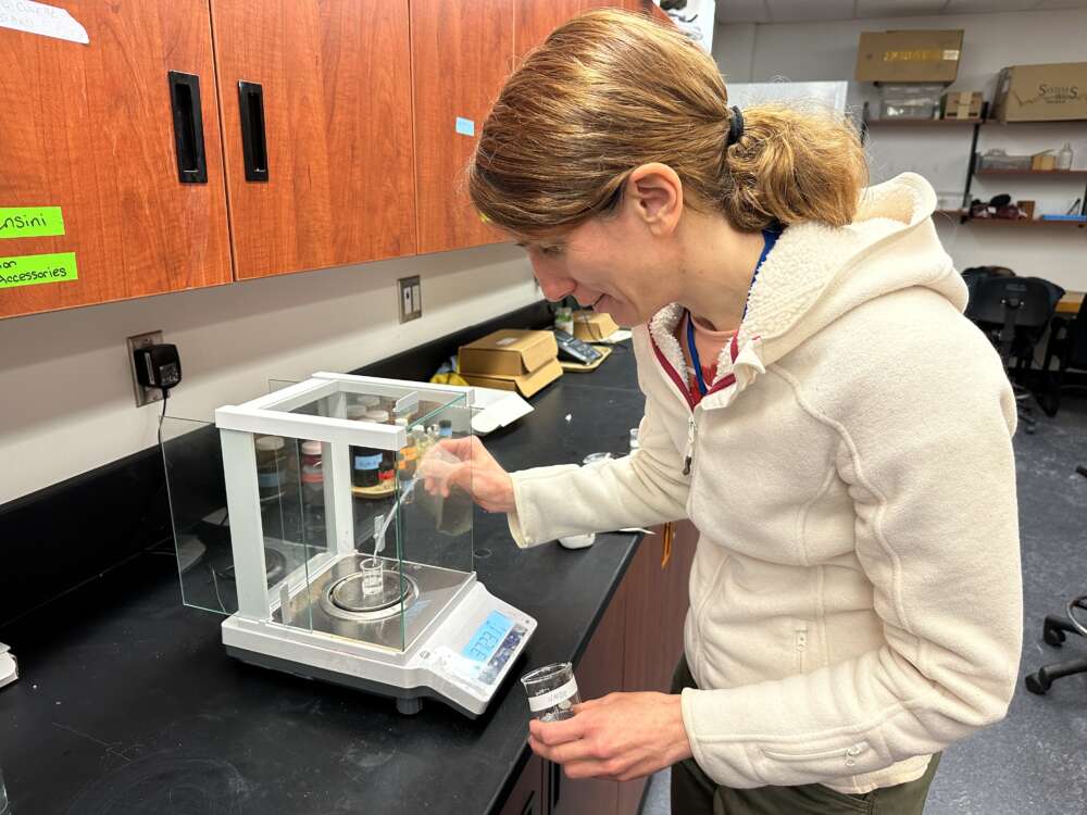 A person wearing a cream-coloured zip-up jacket with a brown ponytail stands in front of lab equipment while holding a small glass jar and tool to create a clear-coloured substance, standing in front of brown cabinets and a black countertop.