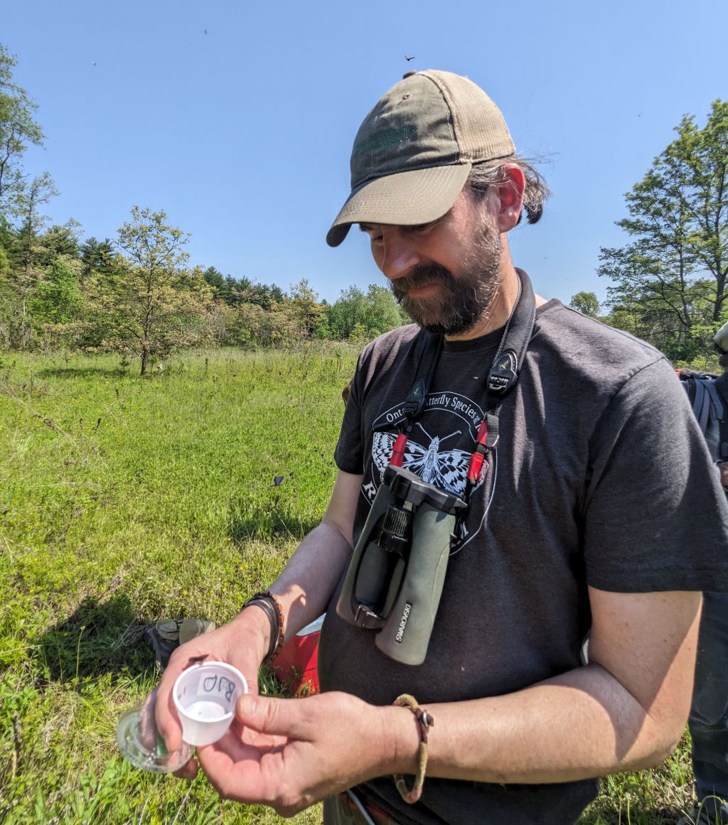a person wearing binoculars around their neck stands in a field and looks down at a specimen container