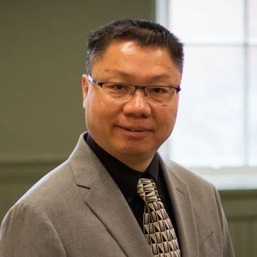 A man with short cropped black hair wearing glasses and a black shirt, beige patterned tie and beige suit jacket smiles into the camera in front of a window.