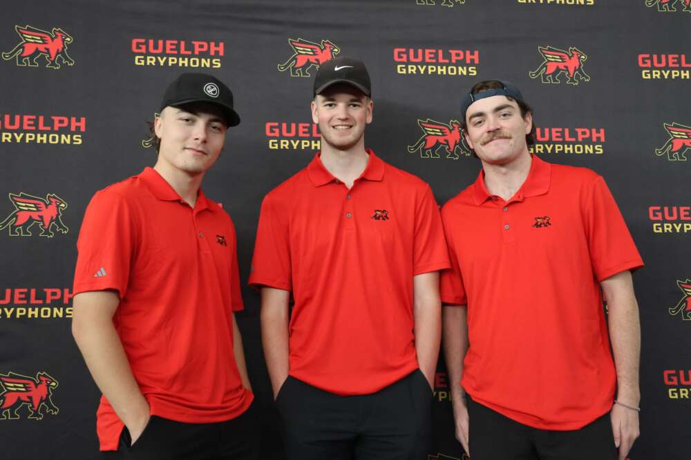 Three people wearing black hats and red U of G golf shirts stand smiling in a row in front of a backdrop with the Guelph Gryphons logo.